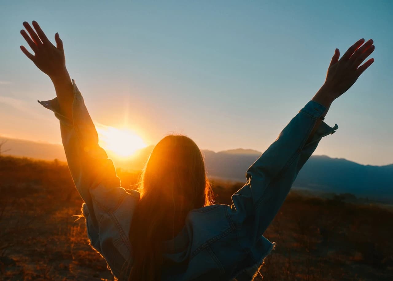 Woman standing on a hillside at sunrise with her arms raised toward the sky — a moment of energised wellbeing